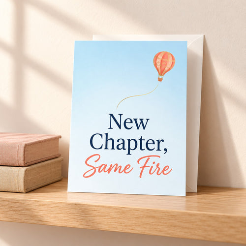 New Chapter, Same Fire card in pale sky blue with a coral hot air balloon, standing on a light oak shelf beside two vintage books