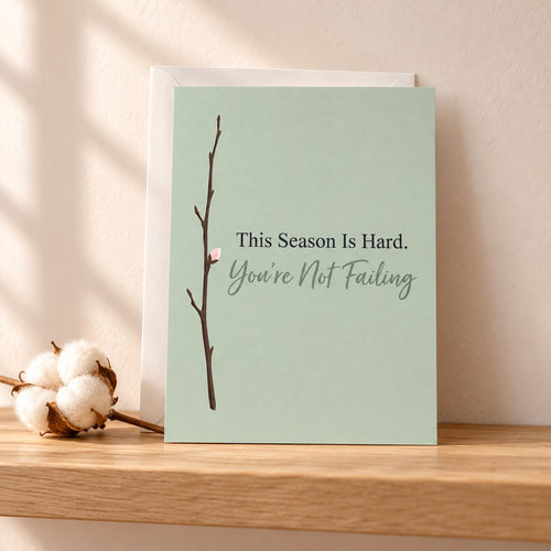 This Season Is Hard. You're Not Failing card in muted sage with a bare twig and one pink bud, standing on a light oak shelf beside a cotton stem