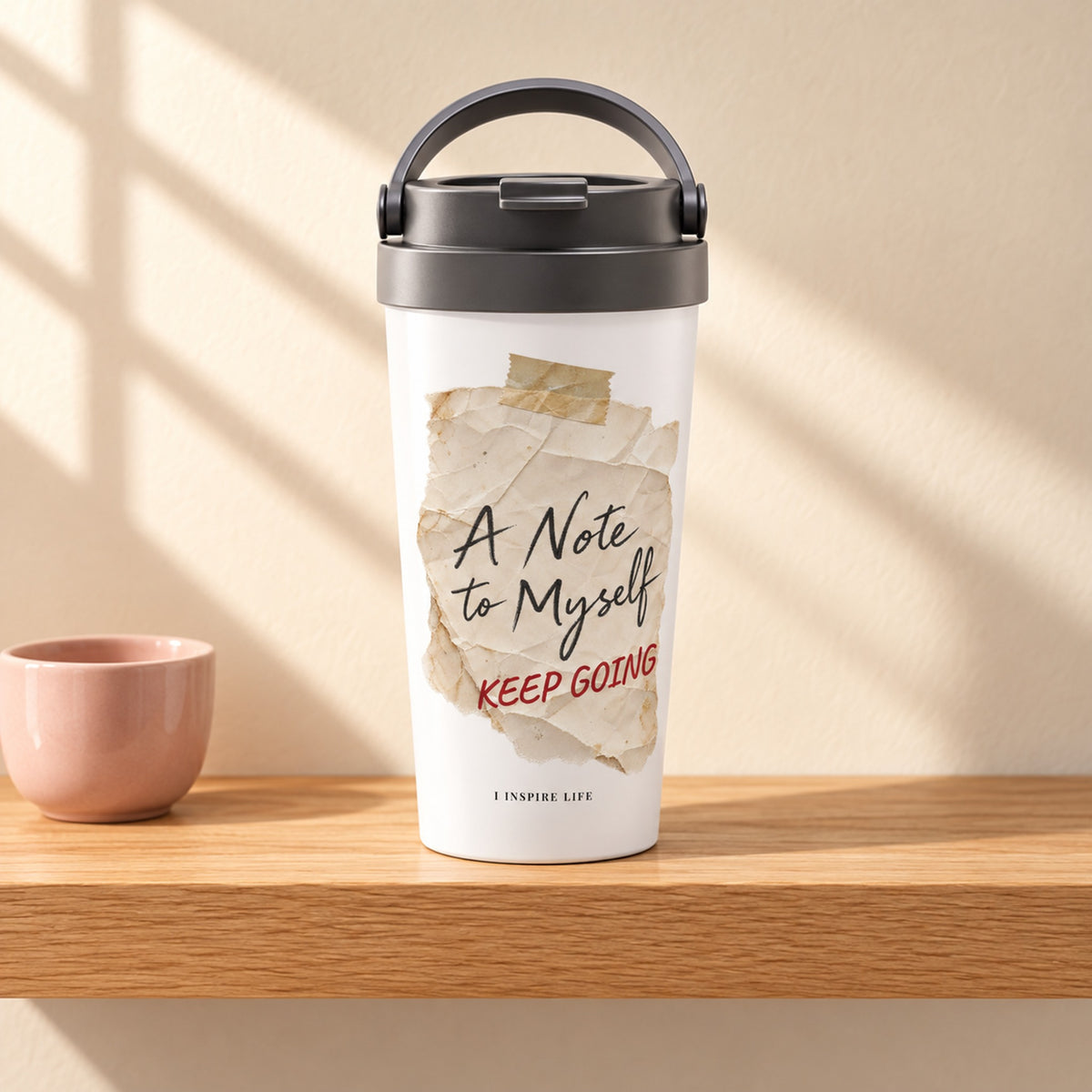 A Note to Myself 15 oz white stainless steel travel mug with KEEP GOING in red, on a light oak shelf beside a small blush pink ceramic cup, design facing forward