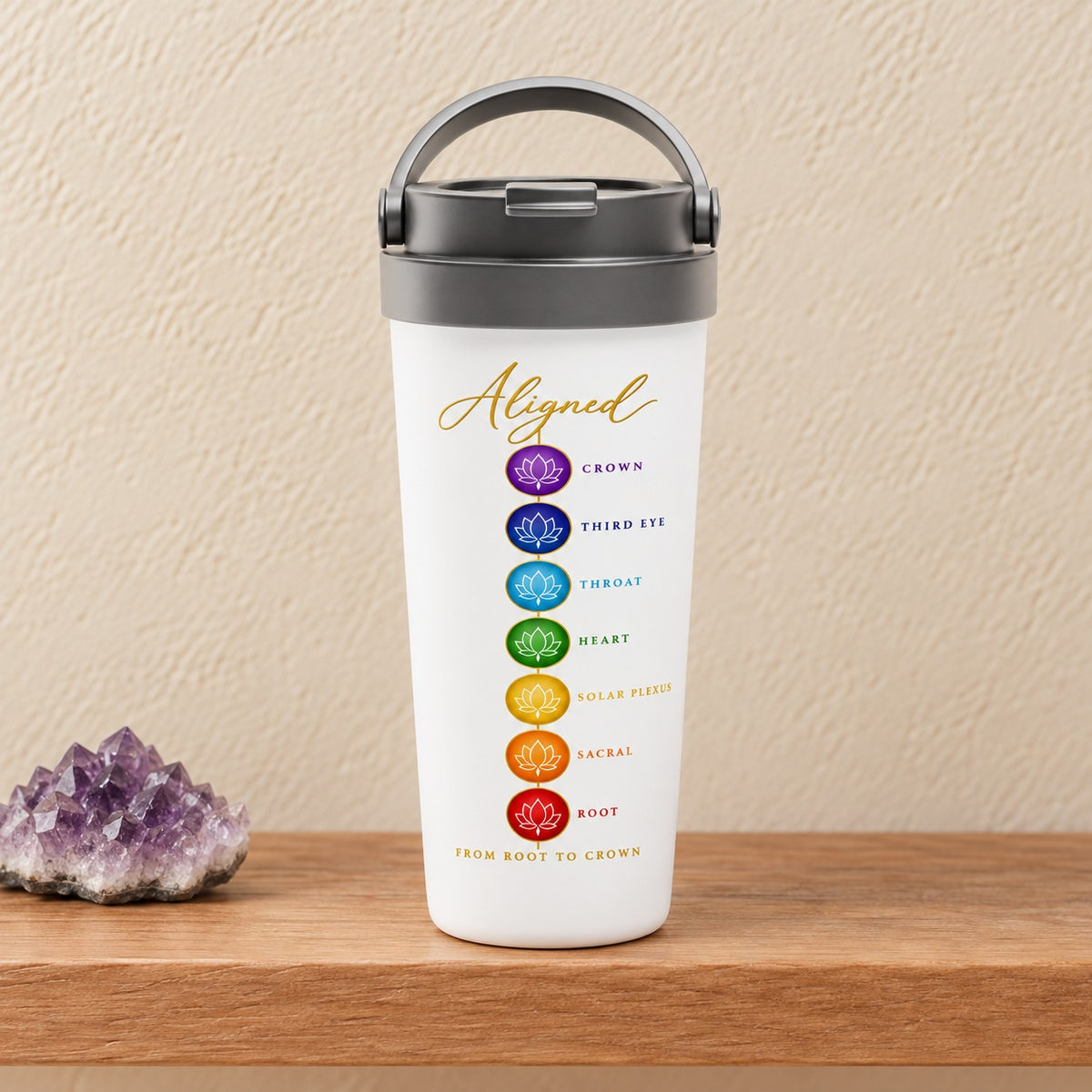 Aligned From Root to Crown 15 oz white travel mug with the seven chakra lotus circles and gold script, standing on a light oak shelf beside an amethyst cluster