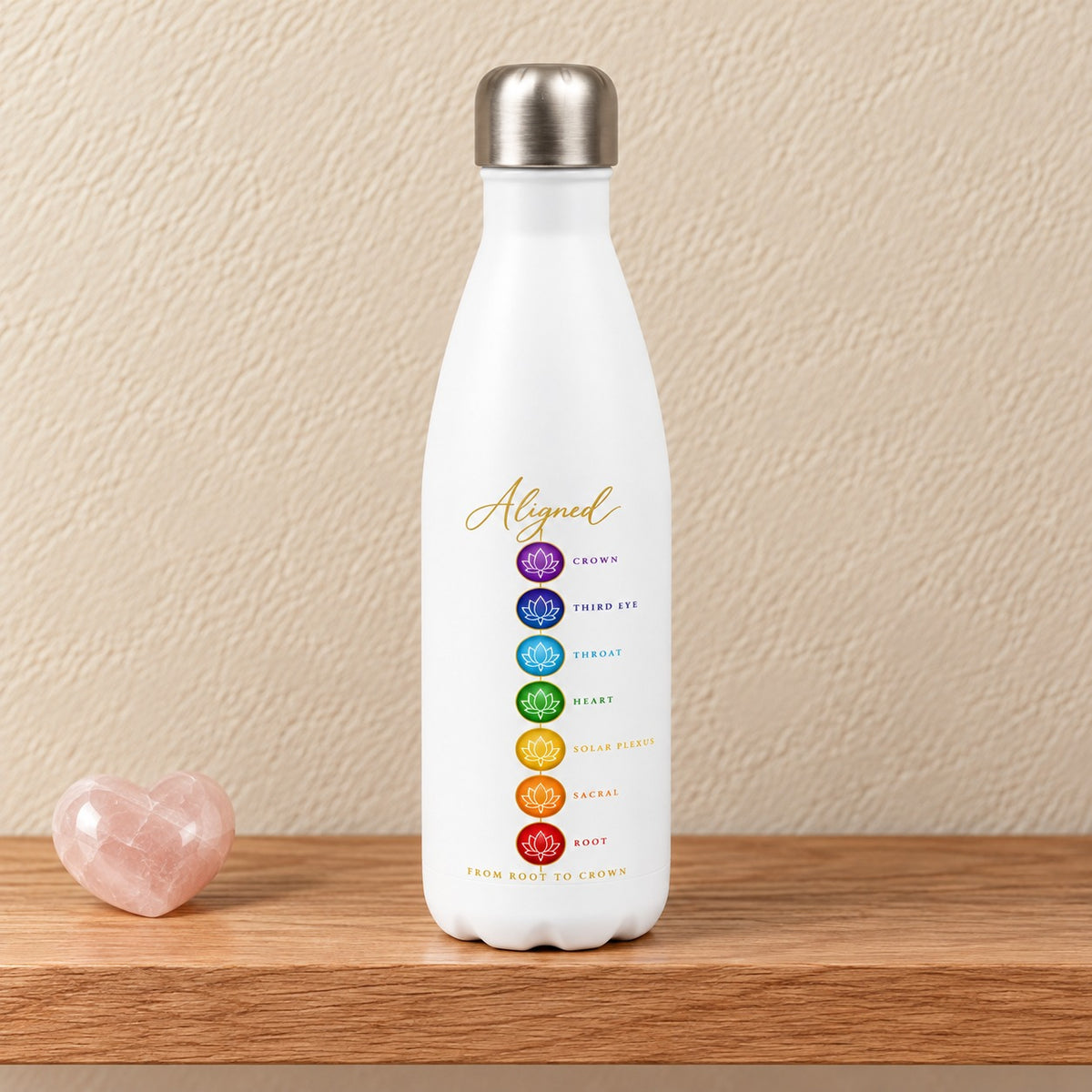 Aligned From Root to Crown 17 oz white water bottle with seven chakra lotus circles and gold script, standing on a light oak shelf beside a rose quartz heart