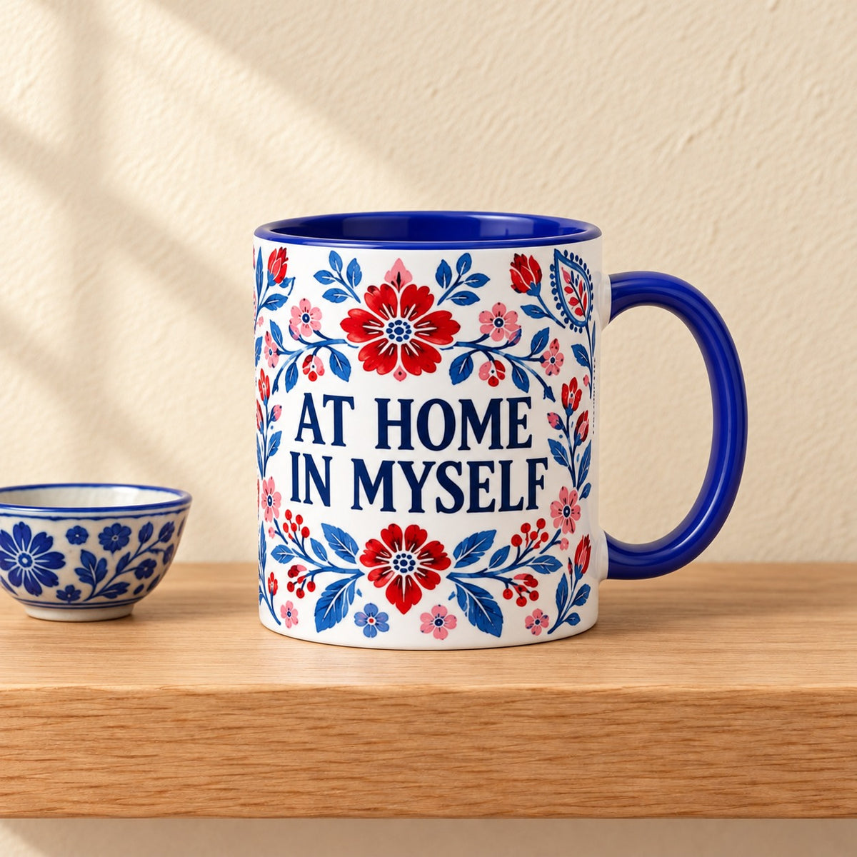 At Home in Myself 11 oz mug with a blue handle and blue inside, title facing forward among blue and red folk florals, resting on a light oak shelf beside a small blue and white bowl