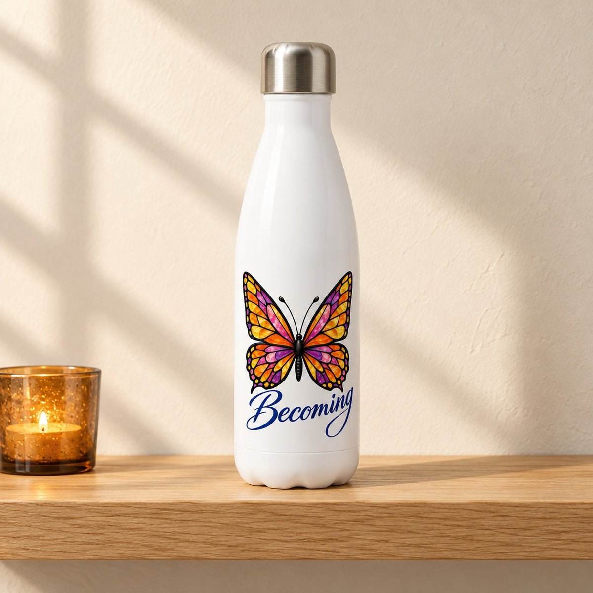 Becoming 17 oz white water bottle with a stained glass butterfly and blue script, standing on a light oak shelf beside an amber candle holder