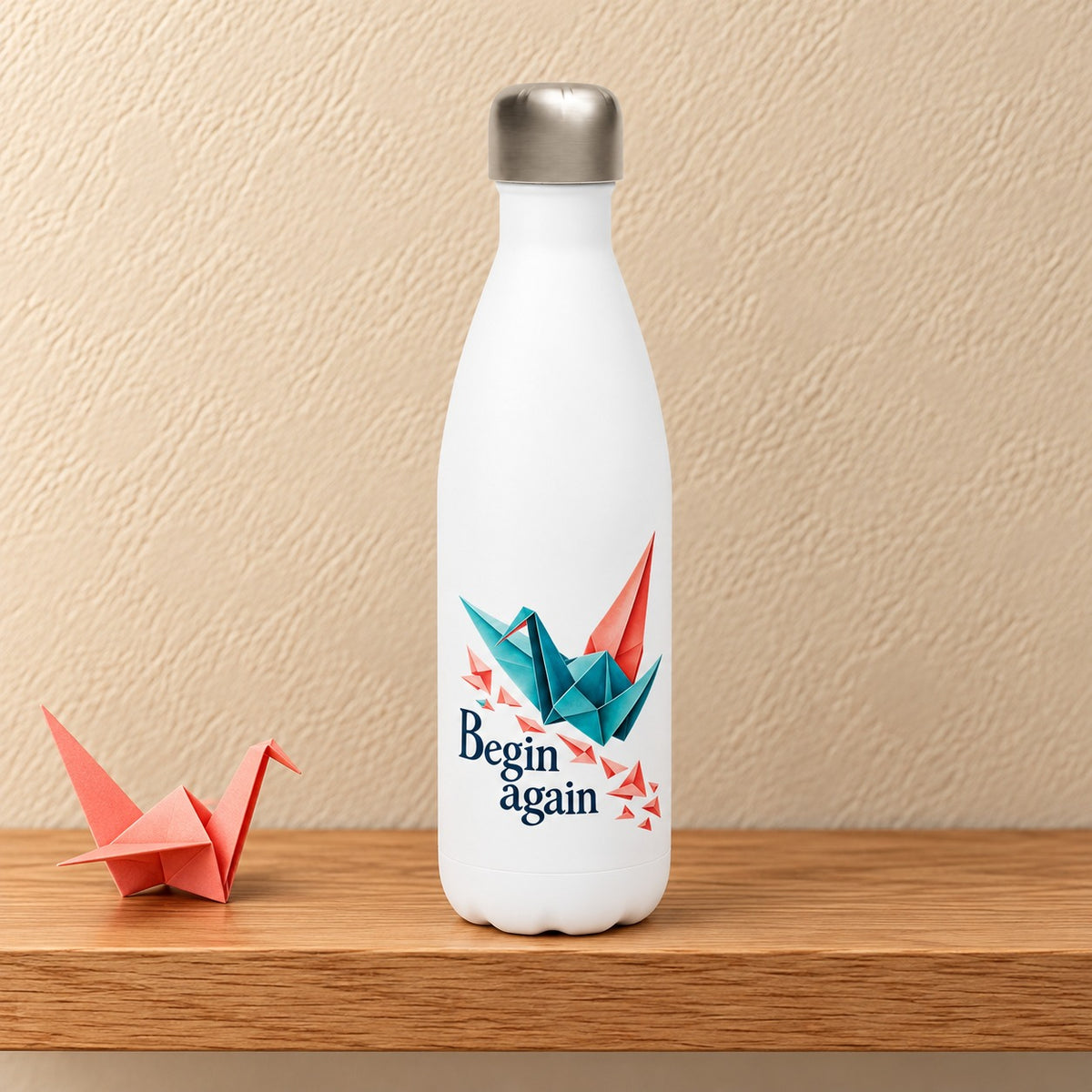 Begin Again 17 oz white water bottle with a teal and coral origami crane, standing on a light oak shelf beside a coral paper crane
