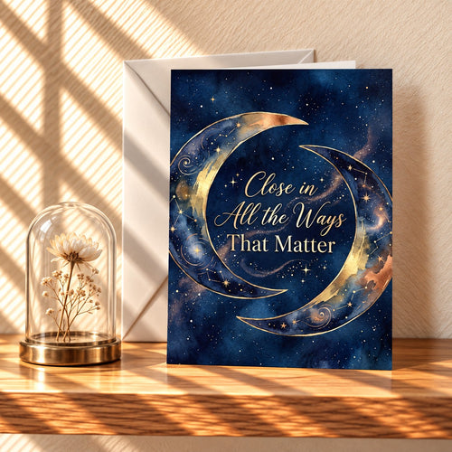 Close in All the Ways That Matter card with two gold crescent moons on a navy starry sky, standing on a light oak shelf beside a glass dome flower