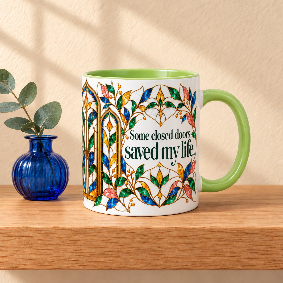 Closed Doors Saved Me 11 oz ceramic mug with a green handle on a light oak shelf beside eucalyptus in a cobalt vase, title facing forward