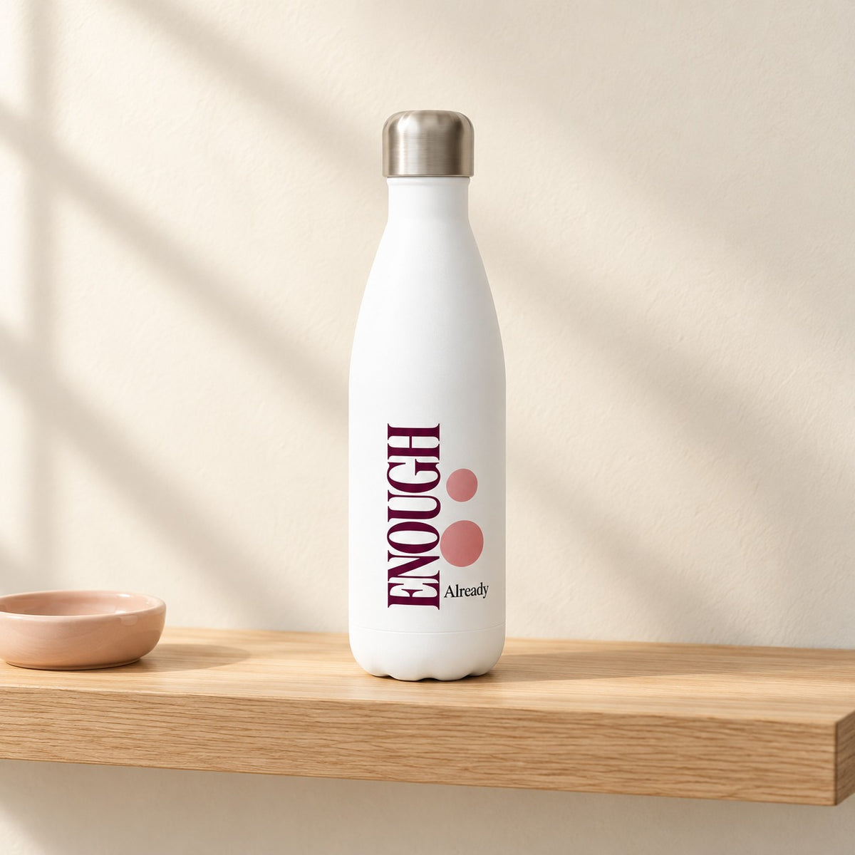 Enough, Already 17 oz white water bottle with a tall plum ENOUGH and pink dots, standing on a light oak shelf beside a blush ring dish