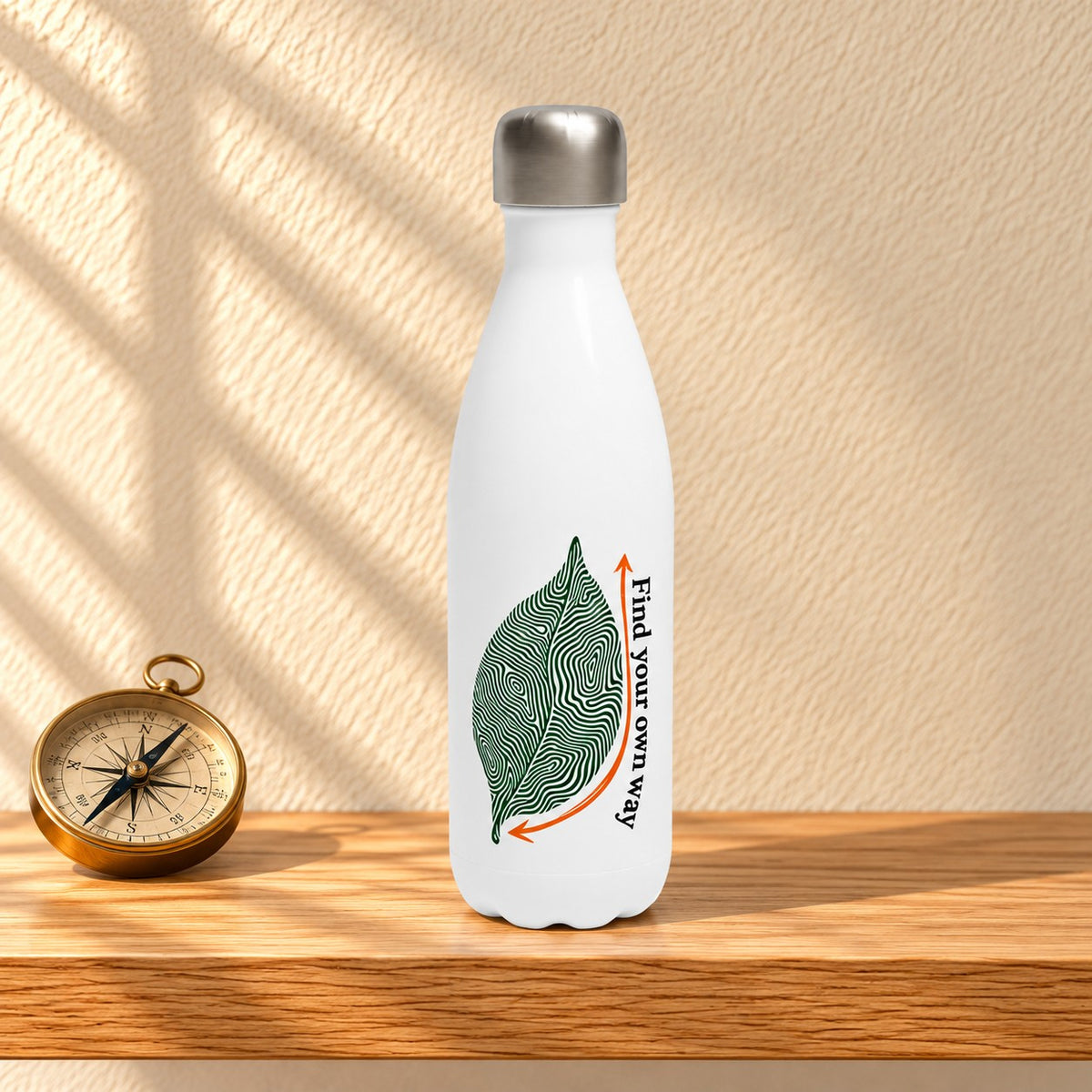 Find Your Own Way white 17 oz water bottle with a green contour line leaf and an orange arrow, standing on a light oak shelf beside a brass compass