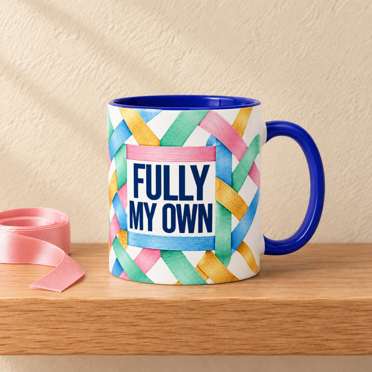 Fully My Own 11 oz mug with a blue handle and blue inside, title facing forward inside woven pastel ribbons, resting on a light oak shelf beside a coil of pink ribbon