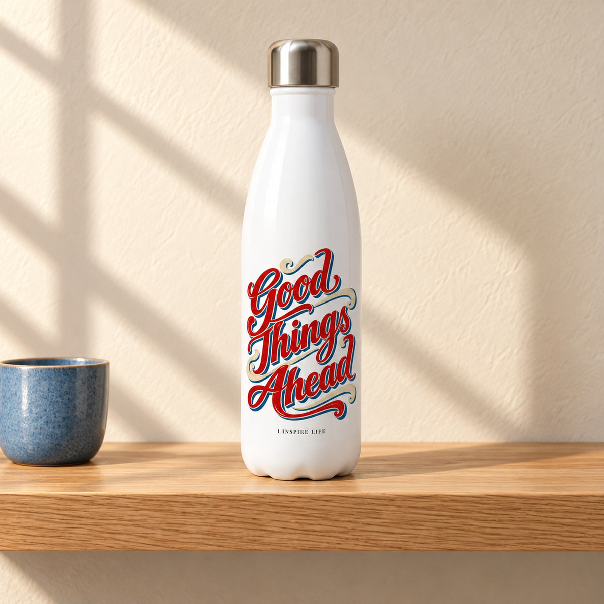 Good Things Ahead 17 oz white stainless steel water bottle on a light oak shelf beside a small blue ceramic cup, design facing forward