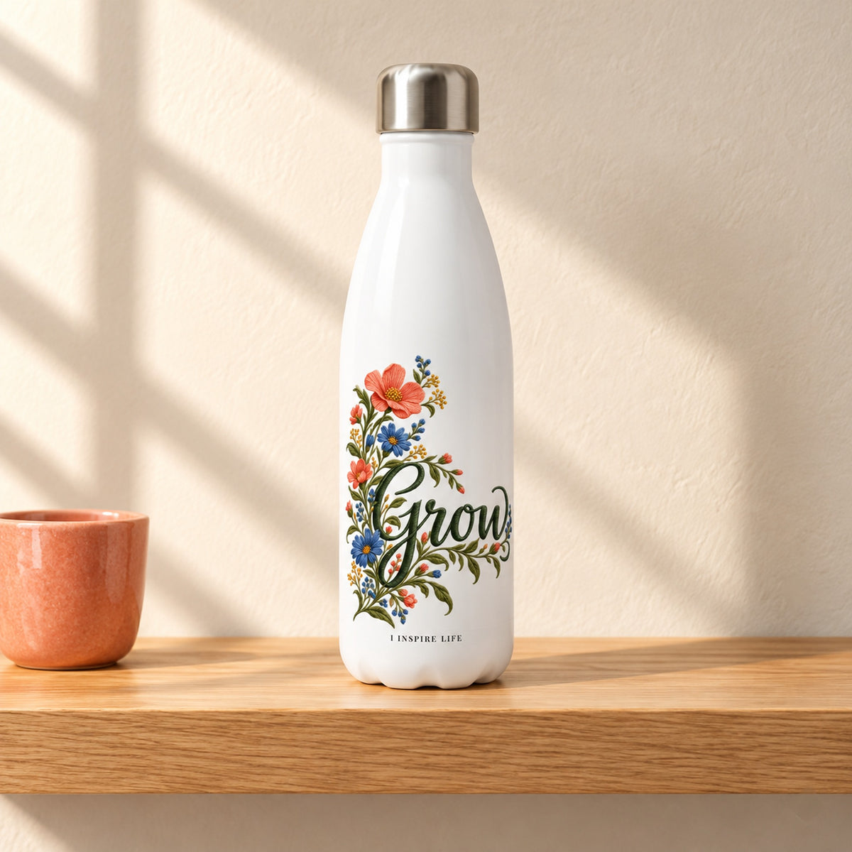 Grow 17 oz white stainless steel water bottle on a light oak shelf beside a small coral ceramic cup, design facing forward