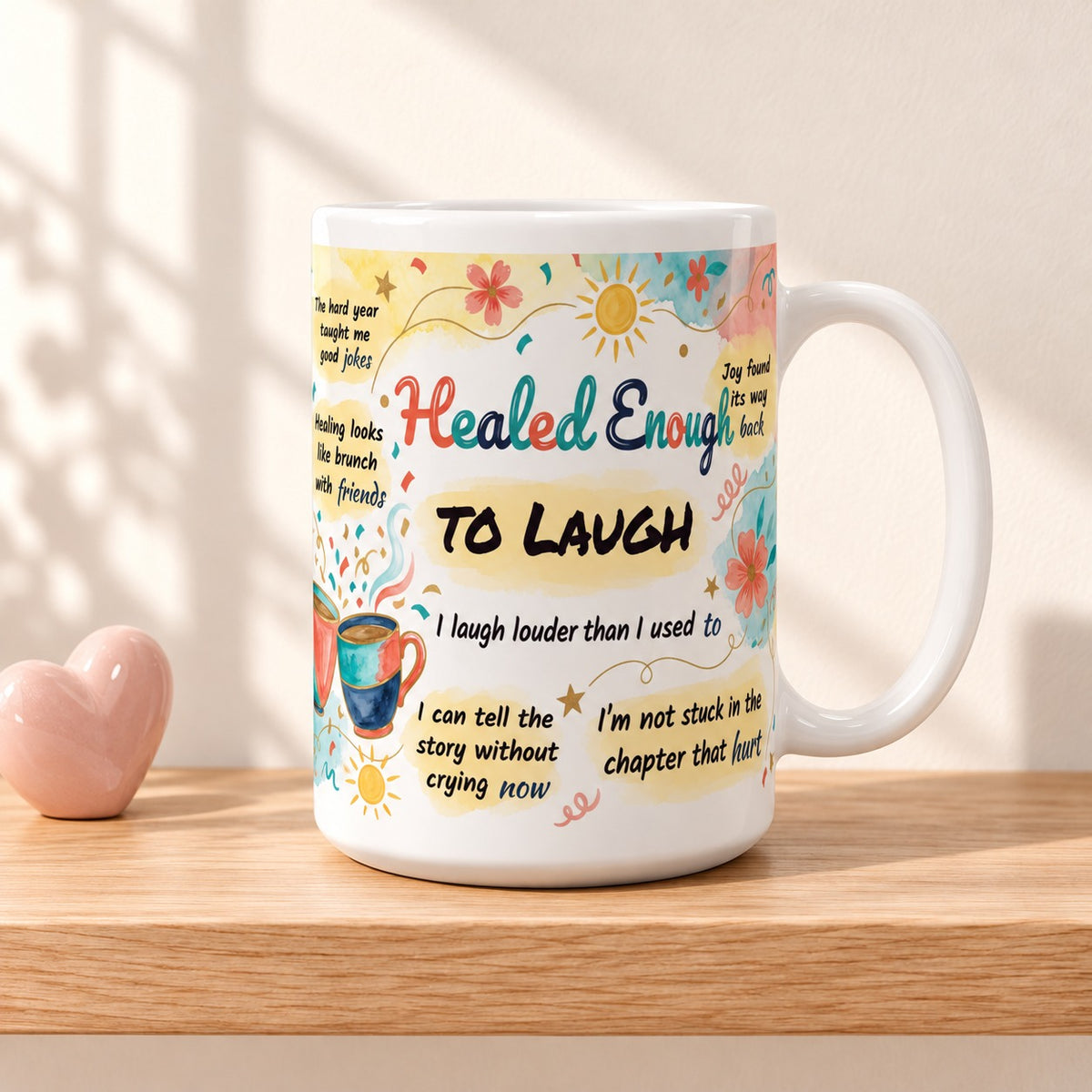 Healed Enough to Laugh 15 oz white mug with the title facing forward, resting on a light oak shelf beside a small pink heart