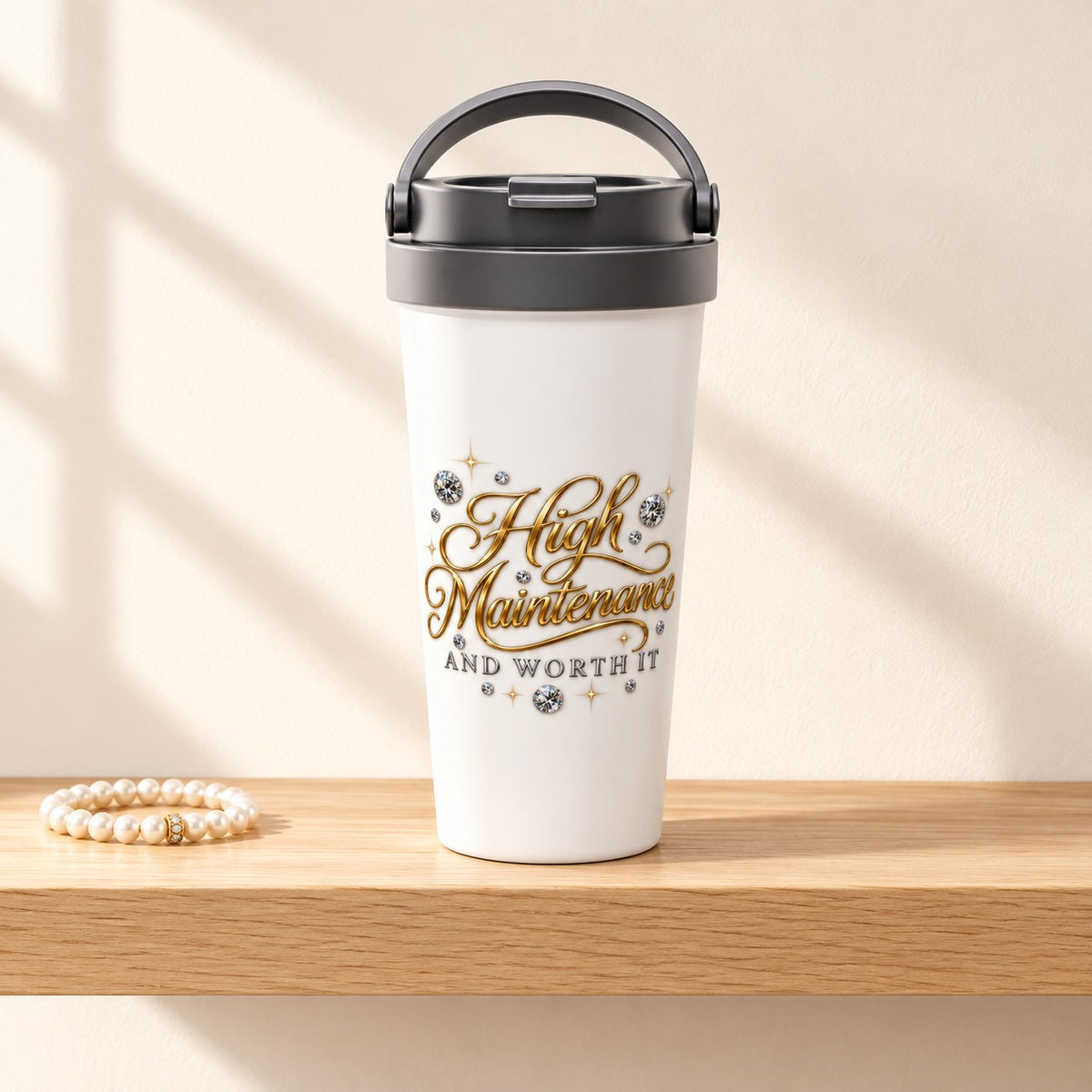 High Maintenance and Worth It 15 oz white travel mug with gold script and diamonds, standing on a light oak shelf beside a pearl bracelet