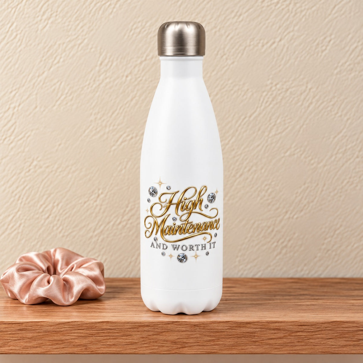 High Maintenance and Worth It 17 oz white water bottle with gold script and diamonds, standing on a light oak shelf beside a blush satin scrunchie