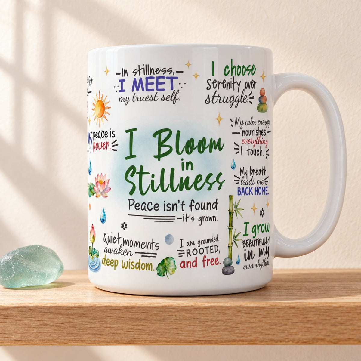 I Bloom in Stillness 15 oz white mug with the green title facing forward among hand lettered affirmations and lotus flowers, on a light oak shelf beside a sea glass stone