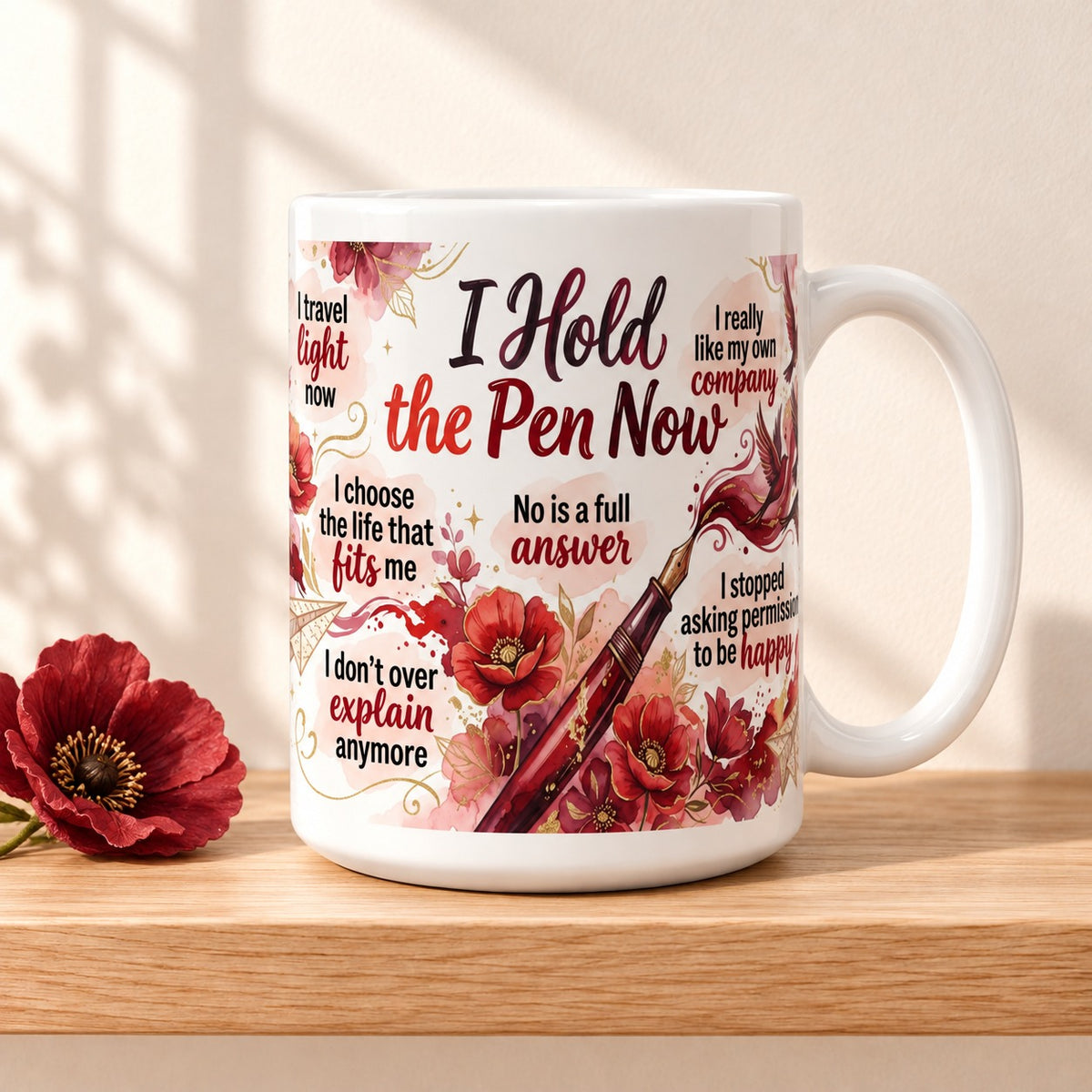 I Hold the Pen Now 15 oz white mug with the title facing forward, resting on a light oak shelf beside a red poppy