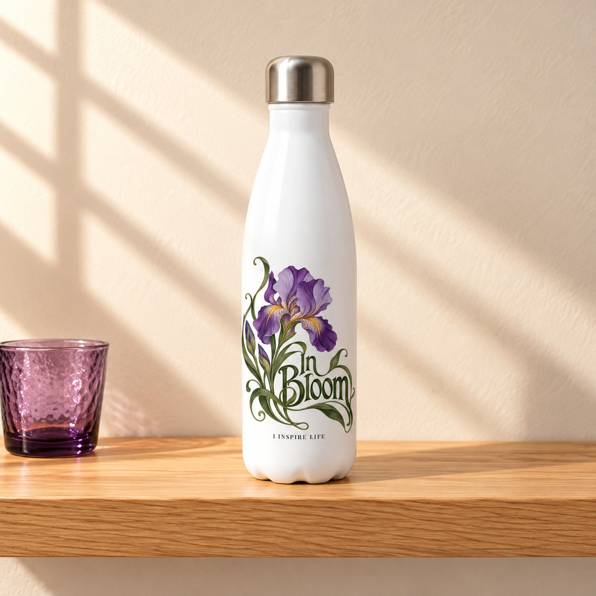 In Bloom 17 oz white stainless steel water bottle on a light oak shelf beside a small purple glass cup, design facing forward
