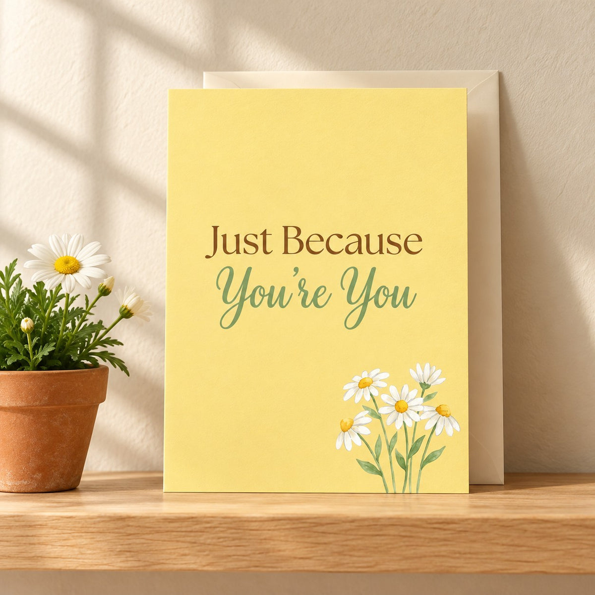 Just Because You're You card in butter yellow with white daisies, standing on a light oak shelf beside a potted daisy