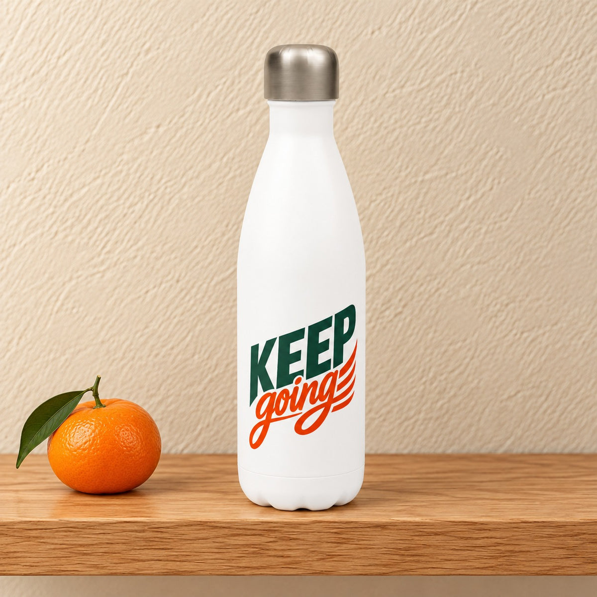Keep Going 17 oz white water bottle with green KEEP and orange going script, standing on a light oak shelf beside a clementine