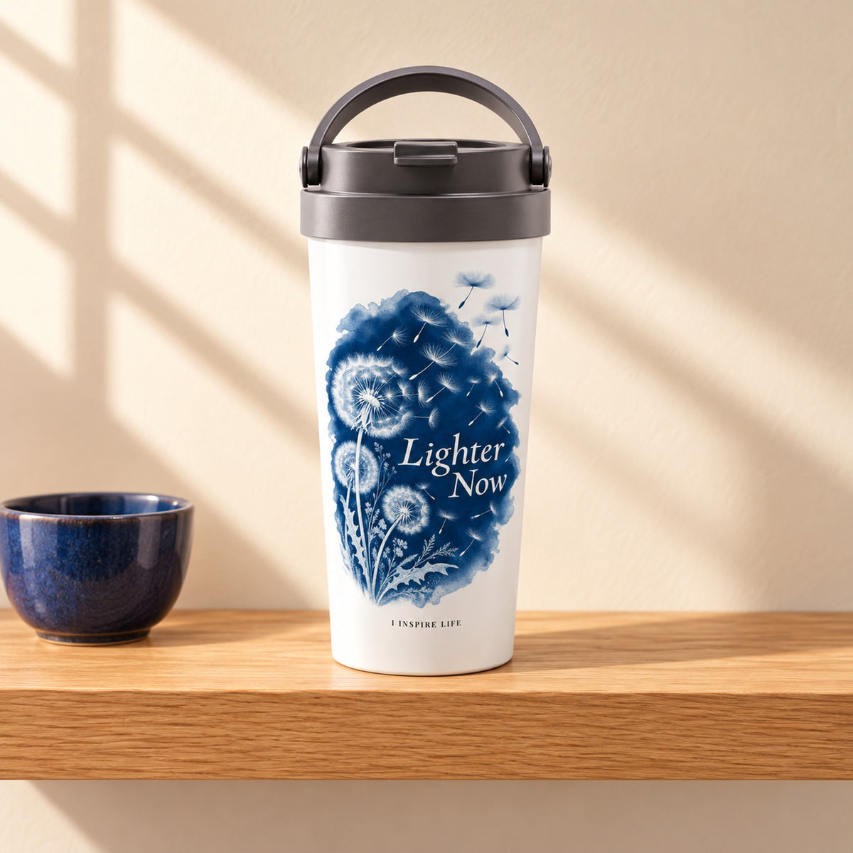 Lighter Now 15 oz white stainless steel travel mug on a light oak shelf beside a small deep blue ceramic cup, design facing forward