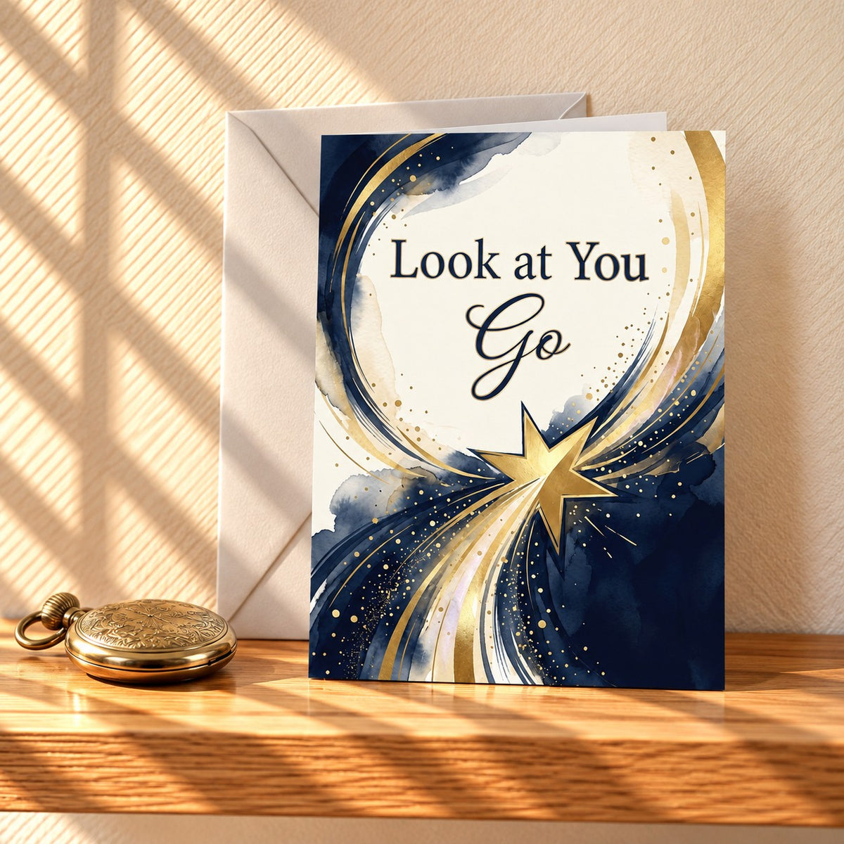 Look at You Go card with a navy and gold watercolour swirl and a gold shooting star, standing on a light oak shelf beside a brass pocket watch