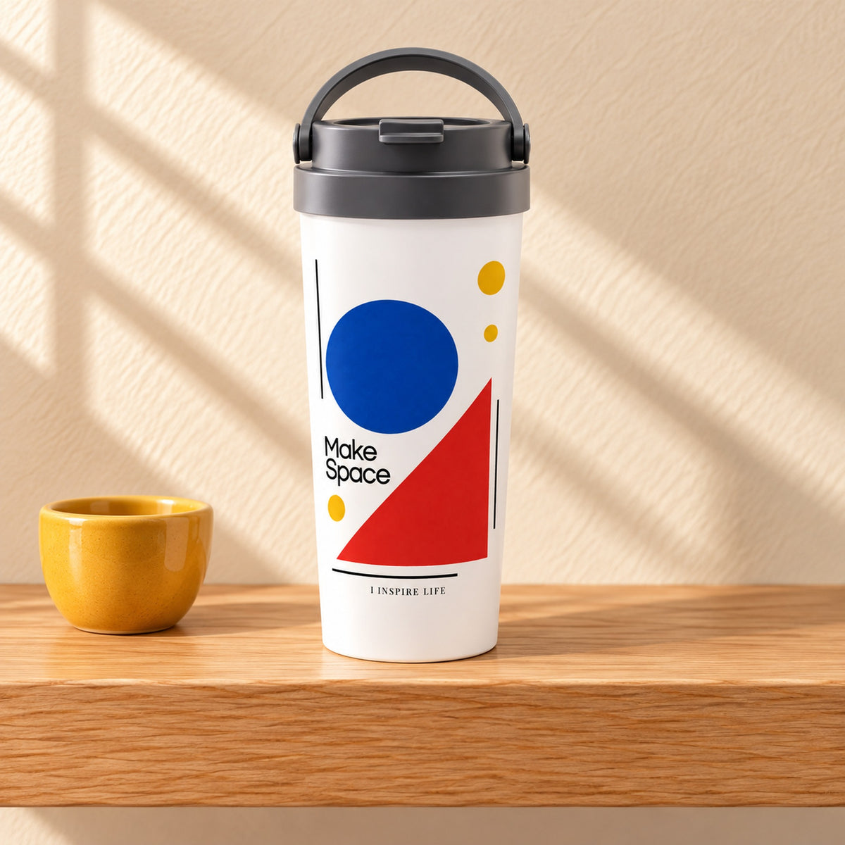 Make Space 15 oz white stainless steel travel mug on a light oak shelf beside a small mustard yellow ceramic cup, design facing forward