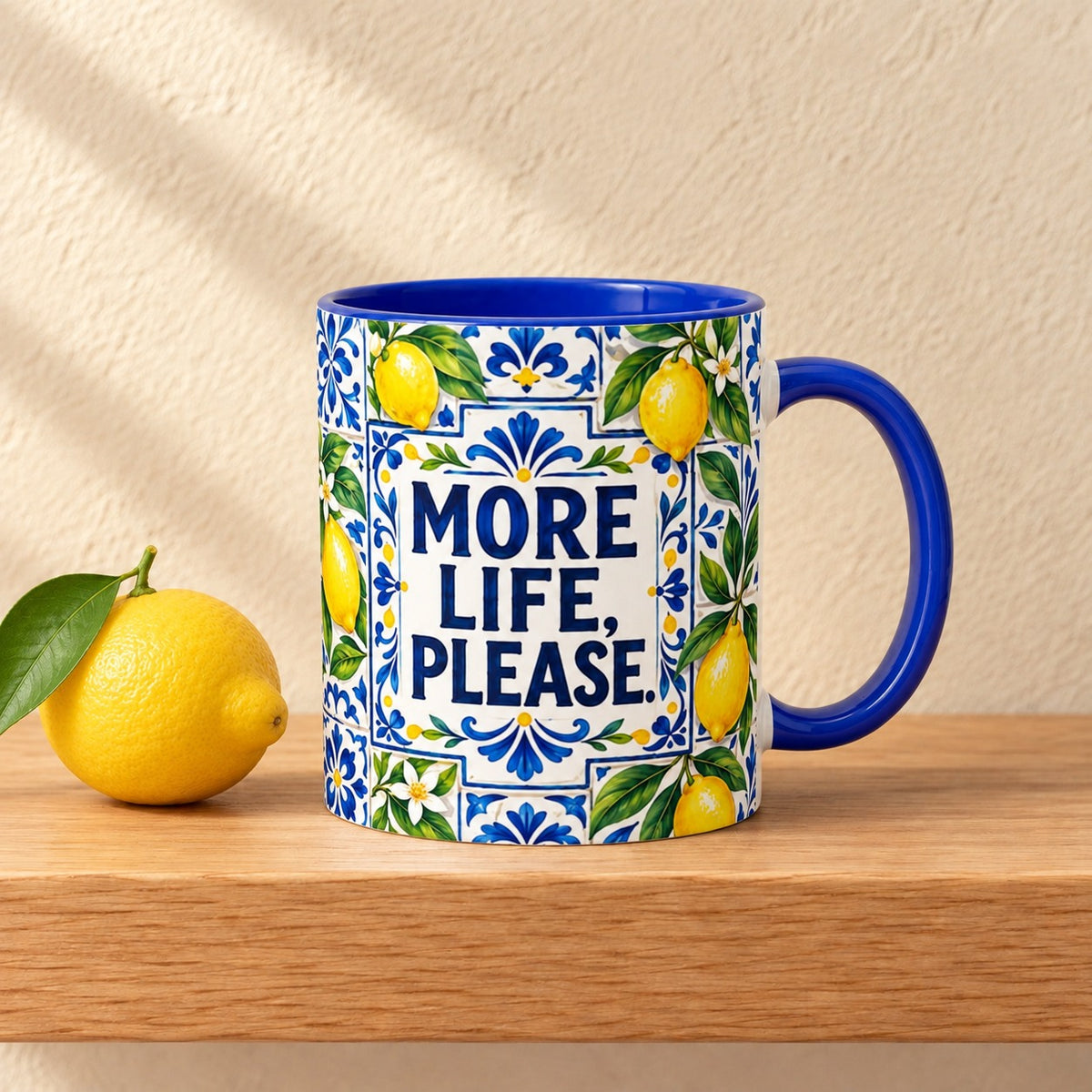More Life, Please 11 oz mug with a blue handle and blue inside, title facing forward among blue tiles and lemons, resting on a light oak shelf beside a fresh lemon