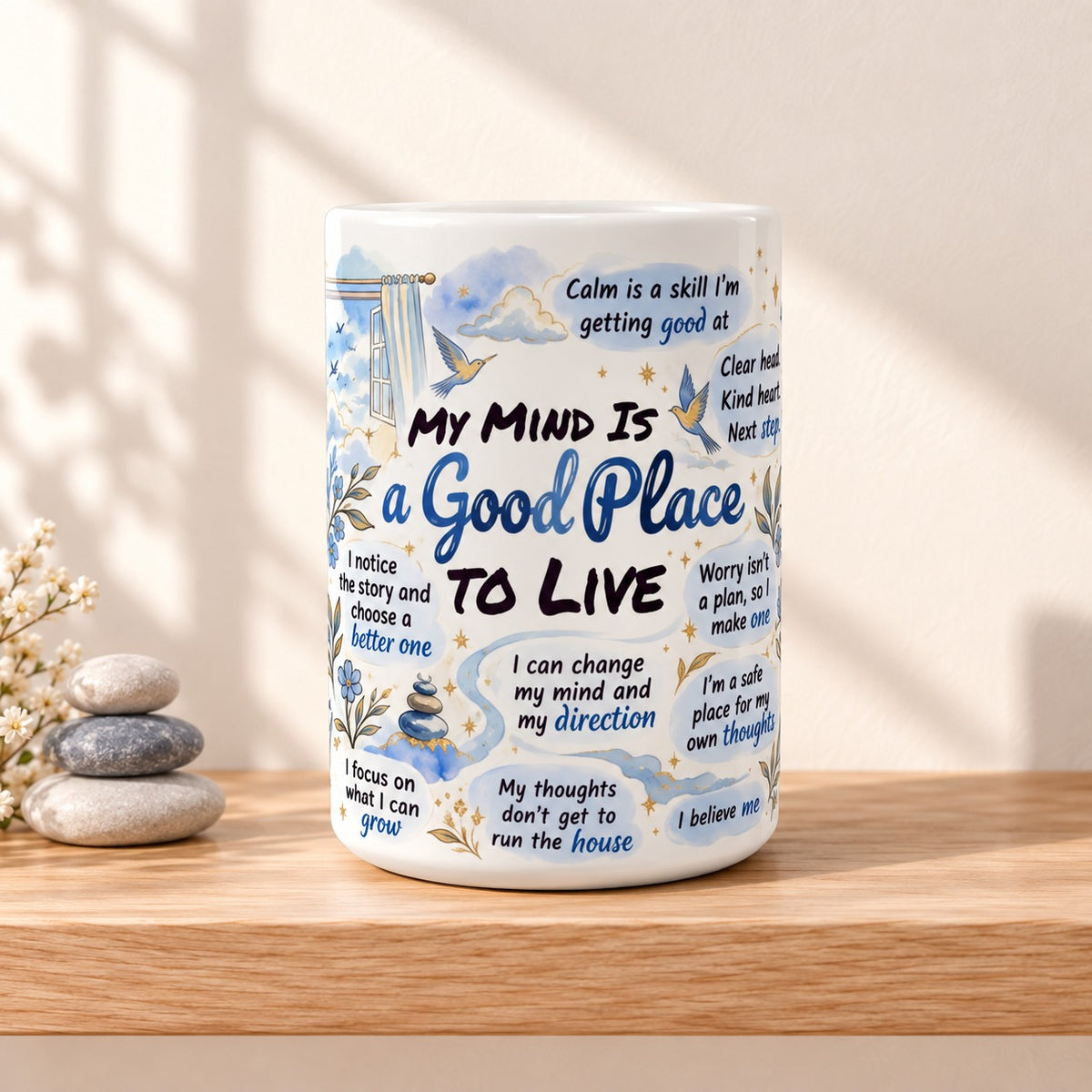 My Mind Is a Good Place to Live 15 oz white mug with the title facing forward, resting on a light oak shelf beside stacked stones