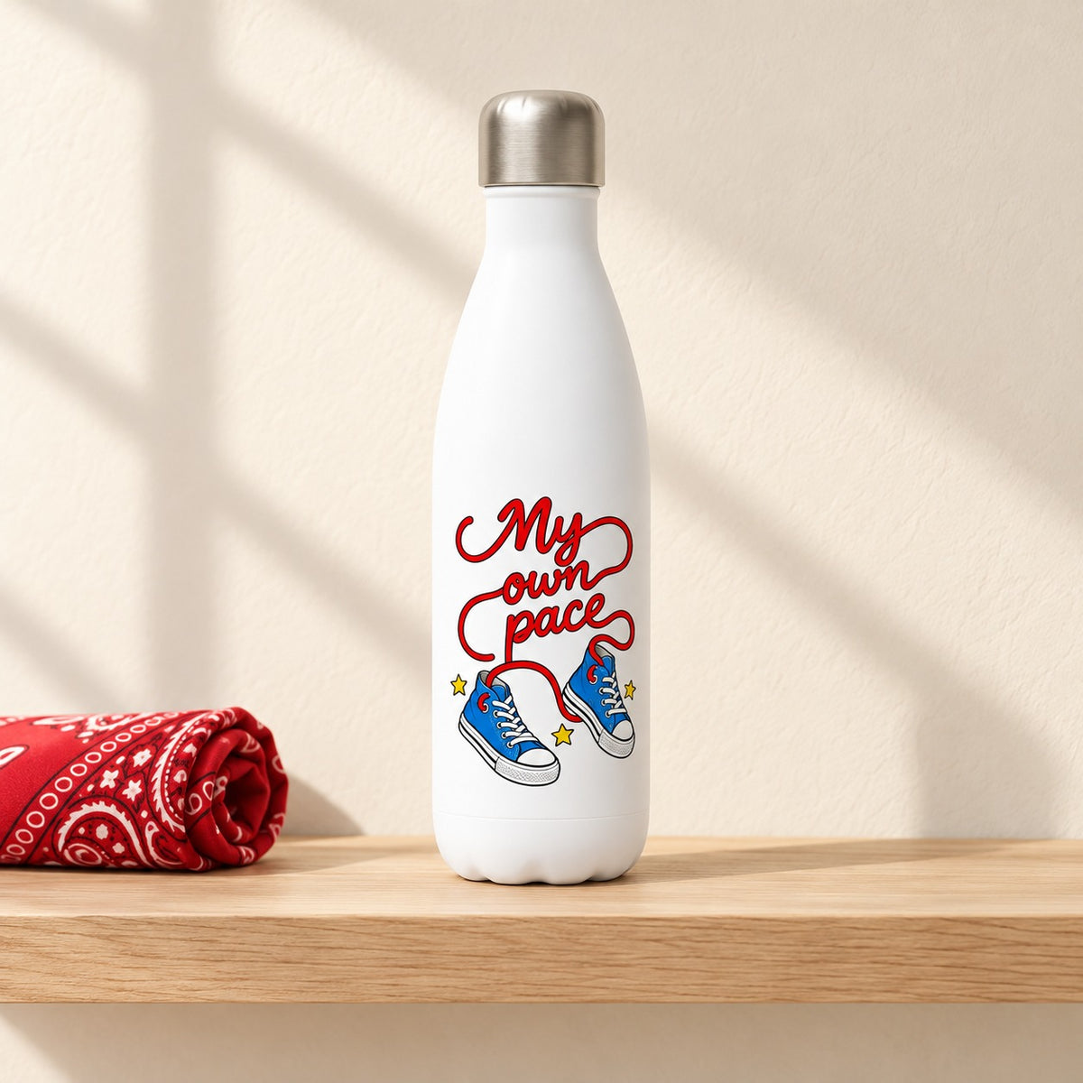 My Own Pace 17 oz white water bottle with red script laces running to blue sneakers, standing on a light oak shelf beside a red bandana