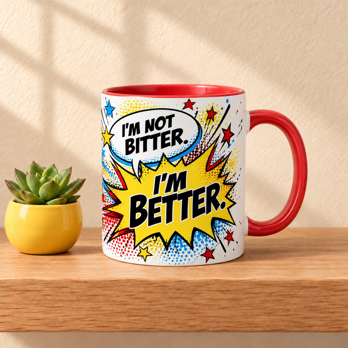 Not Bitter, Better 11 oz ceramic mug with a red handle on a light oak shelf beside a succulent in a yellow pot, title facing forward