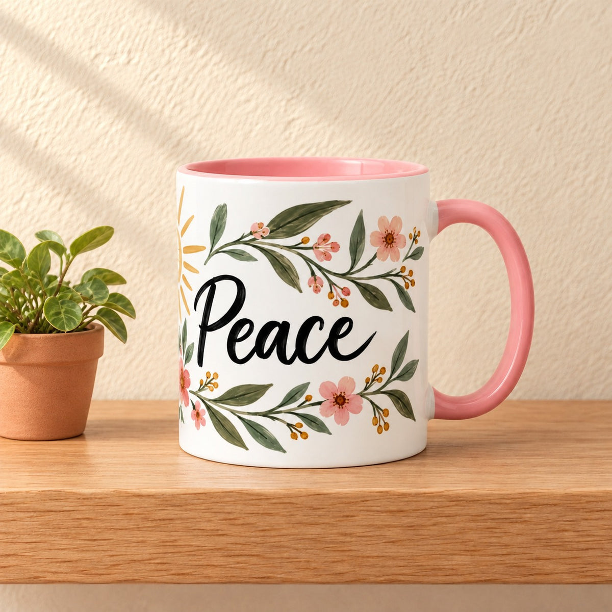Peace 11 oz mug with a pink handle and pink inside, script title facing forward in a soft floral wreath, resting on a light oak shelf beside a small potted plant
