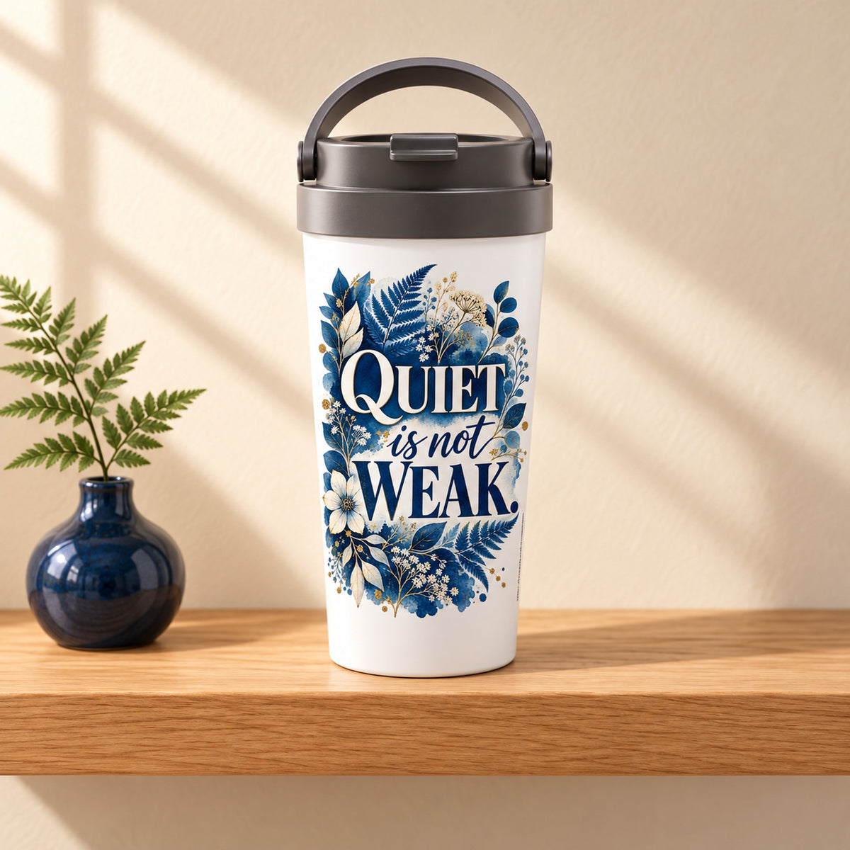 Quiet Is Not Weak 15 oz white stainless steel travel mug on a light oak shelf beside a fern in a navy vase, design facing forward