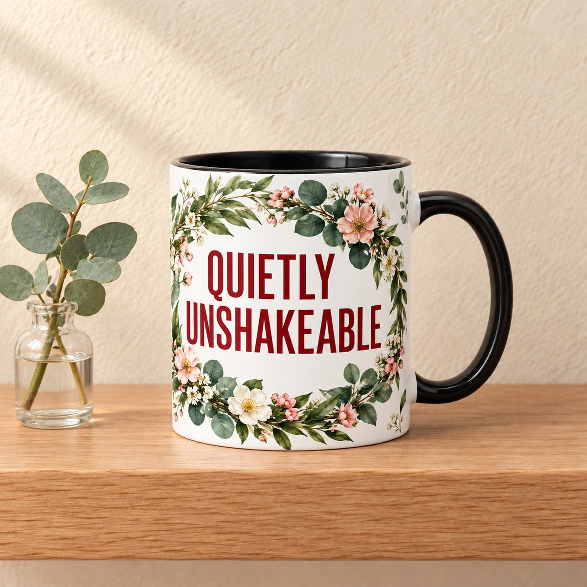 Quietly Unshakeable 11 oz mug with a black handle and black inside, title facing forward inside a blossom and eucalyptus wreath, resting on a light oak shelf beside a eucalyptus sprig