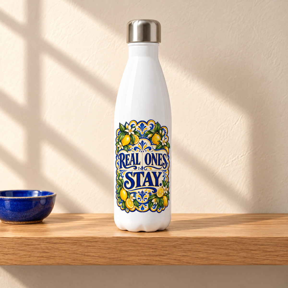 Real Ones Stay 17 oz white stainless steel water bottle on a light oak shelf beside a small cobalt blue ceramic dish, design facing forward