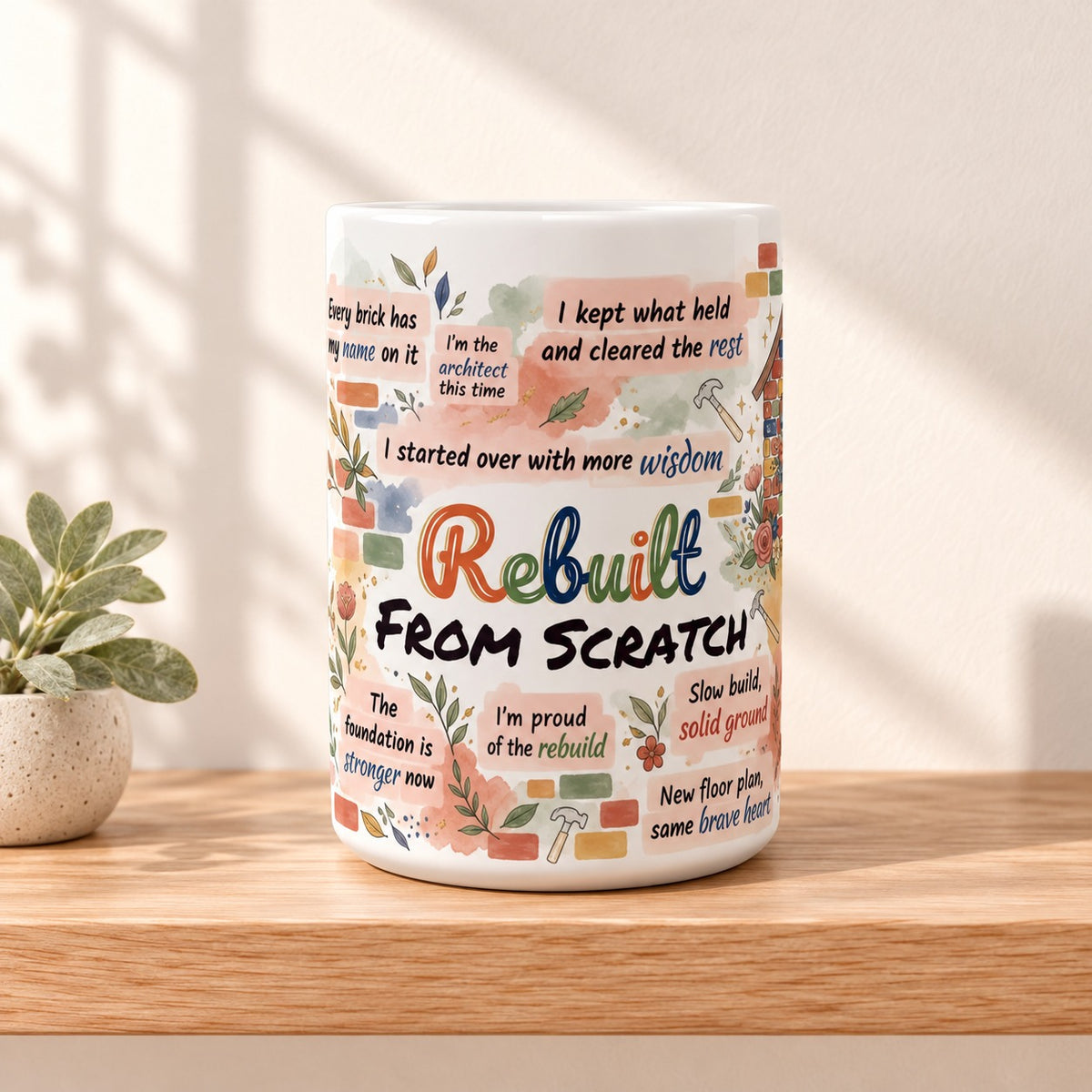Rebuilt From Scratch 15 oz white mug with the title facing forward, resting on a light oak shelf beside a small sage plant