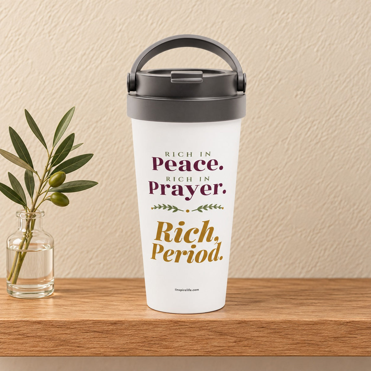 Rich, Period 15 oz white travel mug reading Rich in Peace, Rich in Prayer in plum and gold serif, standing on a light oak shelf beside an olive sprig