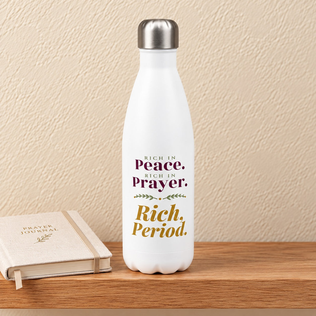 Rich, Period 17 oz white water bottle reading Rich in Peace, Rich in Prayer in plum and gold serif, standing on a light oak shelf beside a linen prayer journal