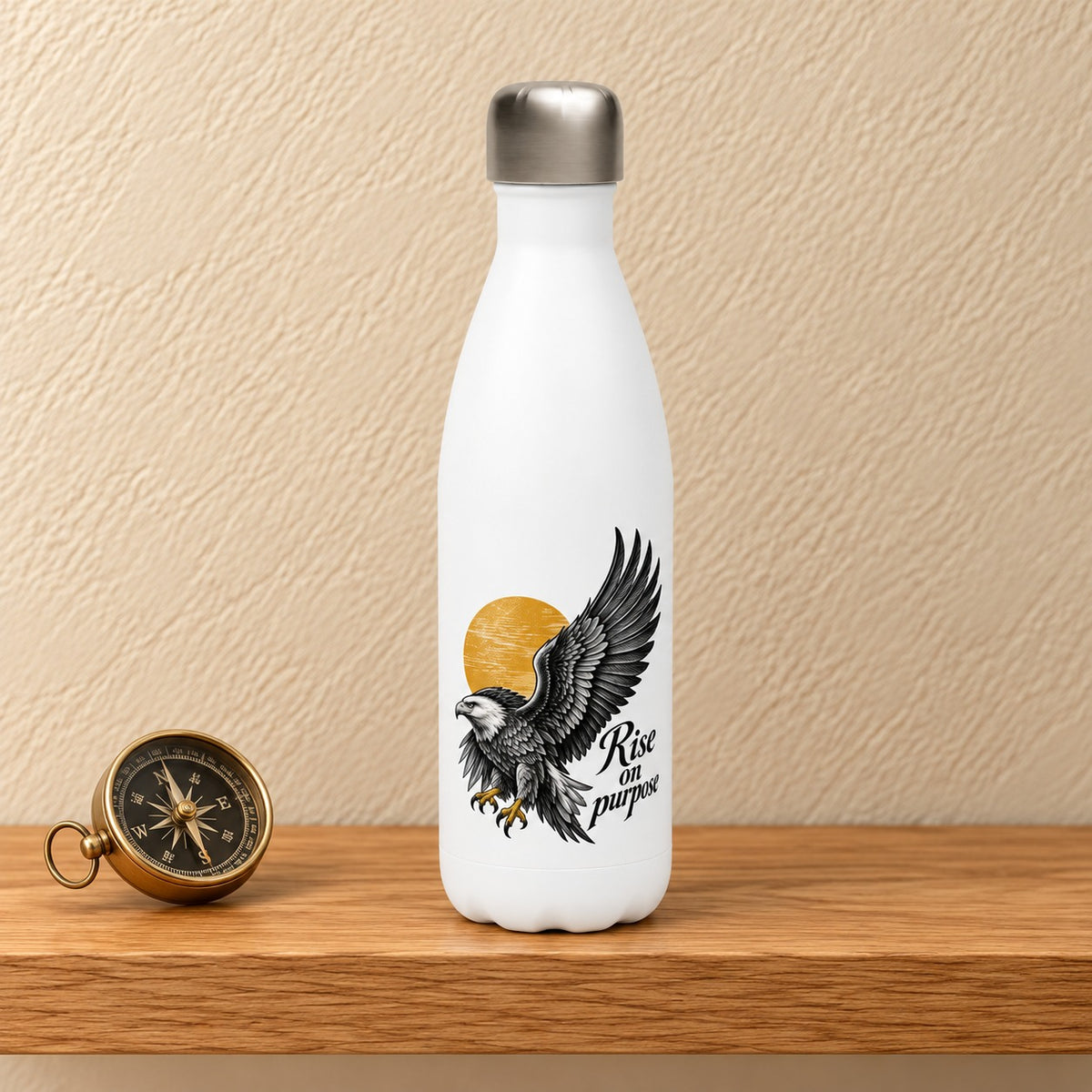 Rise on Purpose 17 oz white water bottle with a black eagle in flight over a gold sun, standing on a light oak shelf beside a brass compass