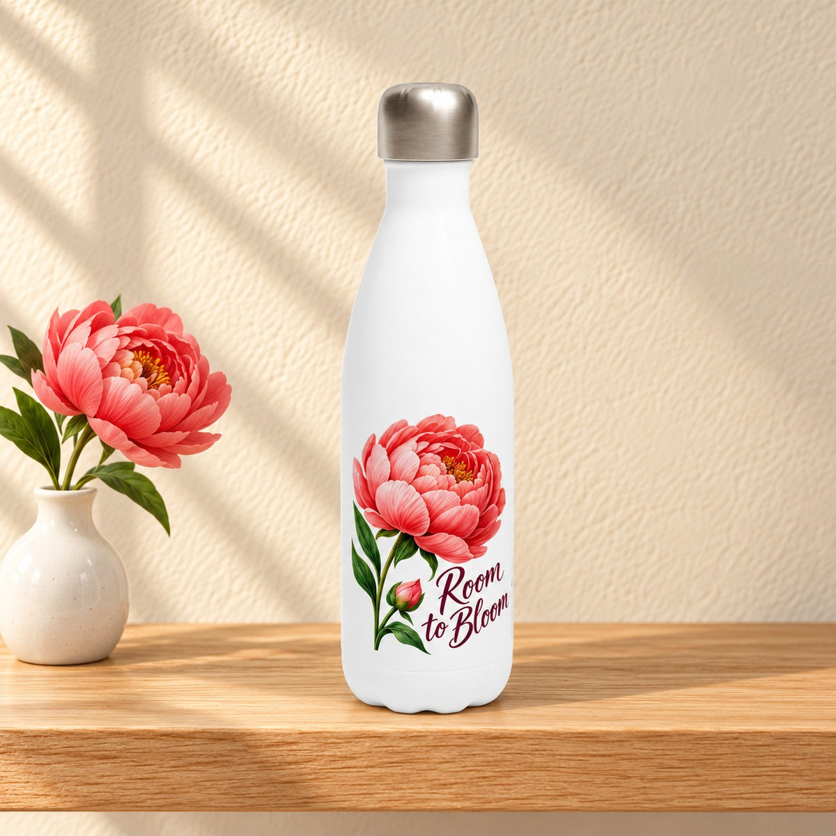 Room to Bloom white 17 oz water bottle with a coral peony and plum script, standing on a light oak shelf beside a peony in a white bud vase