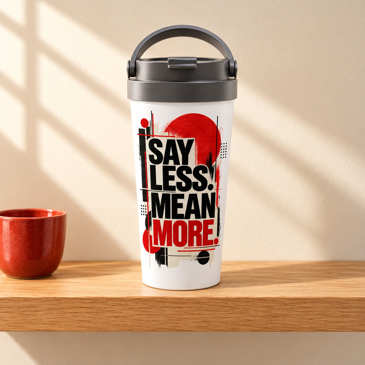 Say Less, Mean More 15 oz white stainless steel travel mug on a light oak shelf beside a red ceramic cup, design facing forward