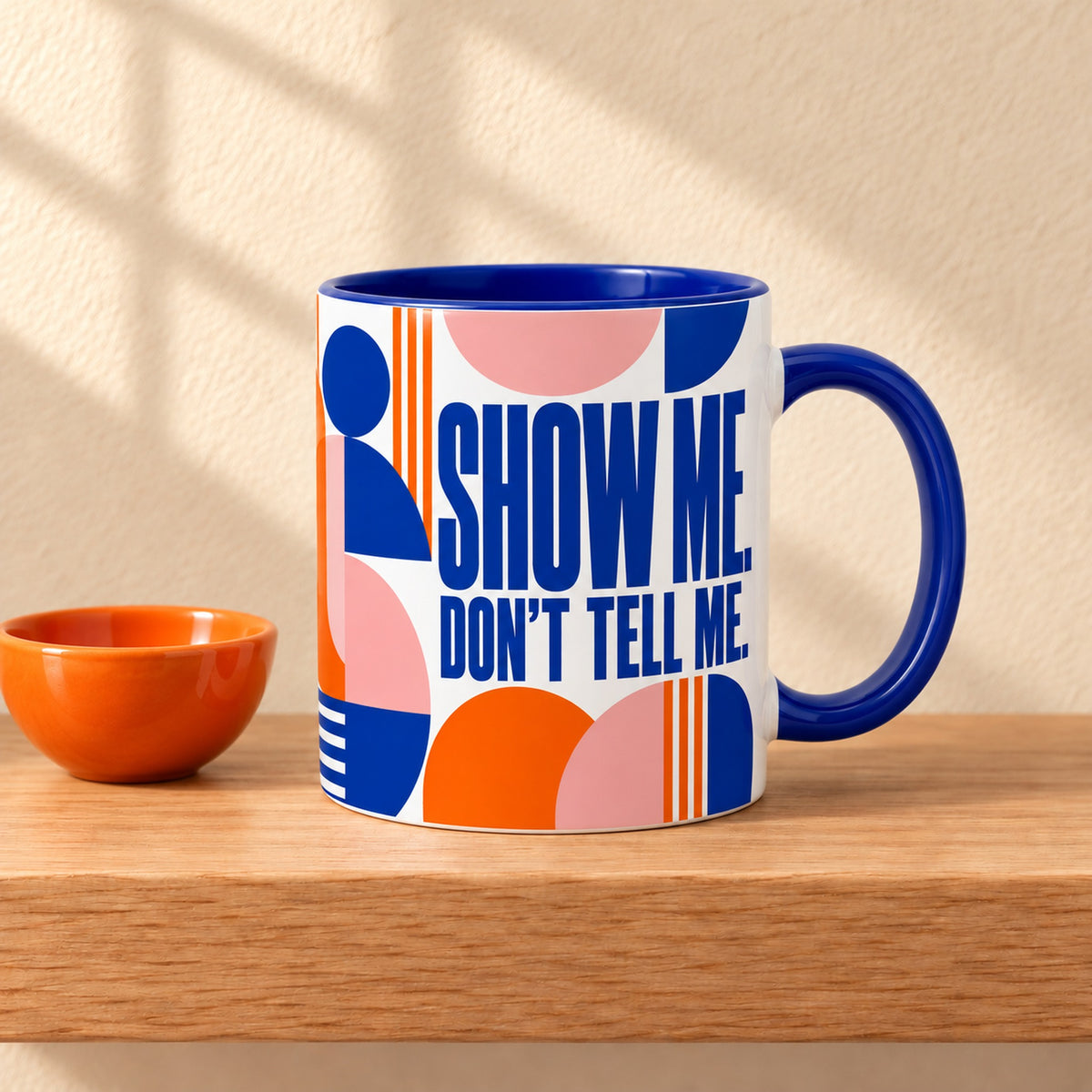 Show Me, Don't Tell Me 11 oz ceramic mug with a blue handle on a light oak shelf beside a small orange bowl, title facing forward