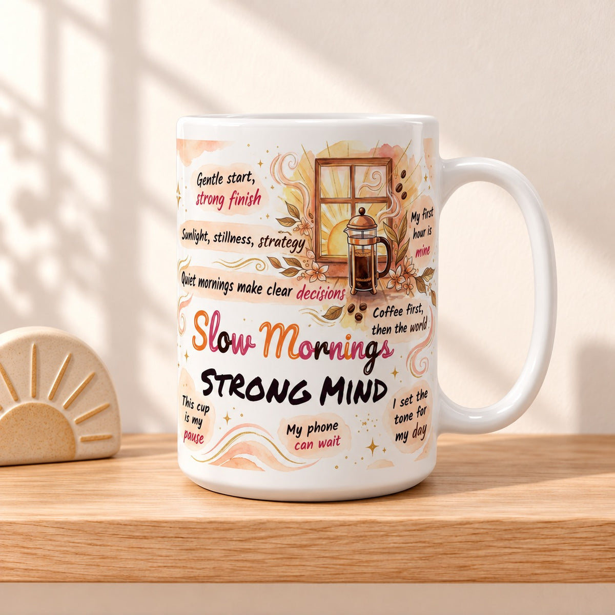 Slow Mornings Strong Mind 15 oz white mug with the title facing forward, resting on a light oak shelf beside a small sunrise ornament