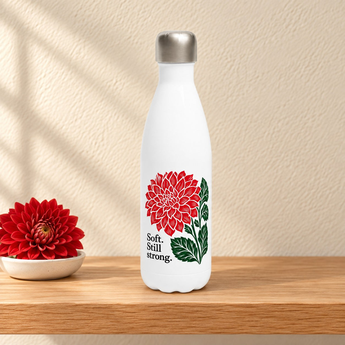 Soft, Still Strong white 17 oz water bottle with a red block print dahlia, standing on a light oak shelf beside a red dahlia in a small dish