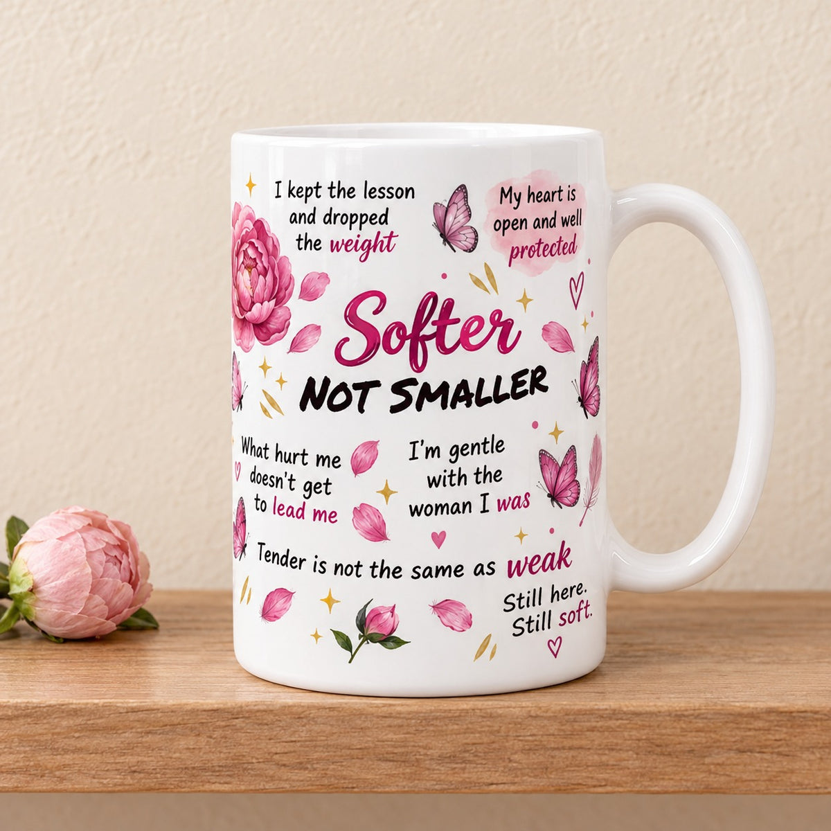 Softer Not Smaller 15 oz white mug with the pink and black title facing forward among gentle affirmations, peonies and butterflies, on a light oak shelf beside a blush peony bud