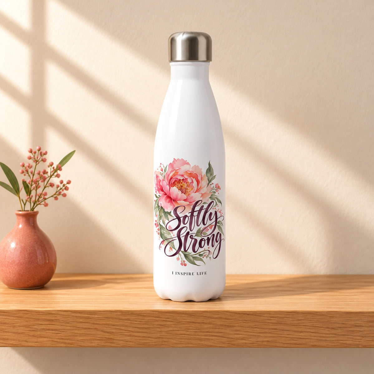 Softly Strong 17 oz white stainless steel water bottle on a light oak shelf beside a small coral ceramic bud vase, design facing forward