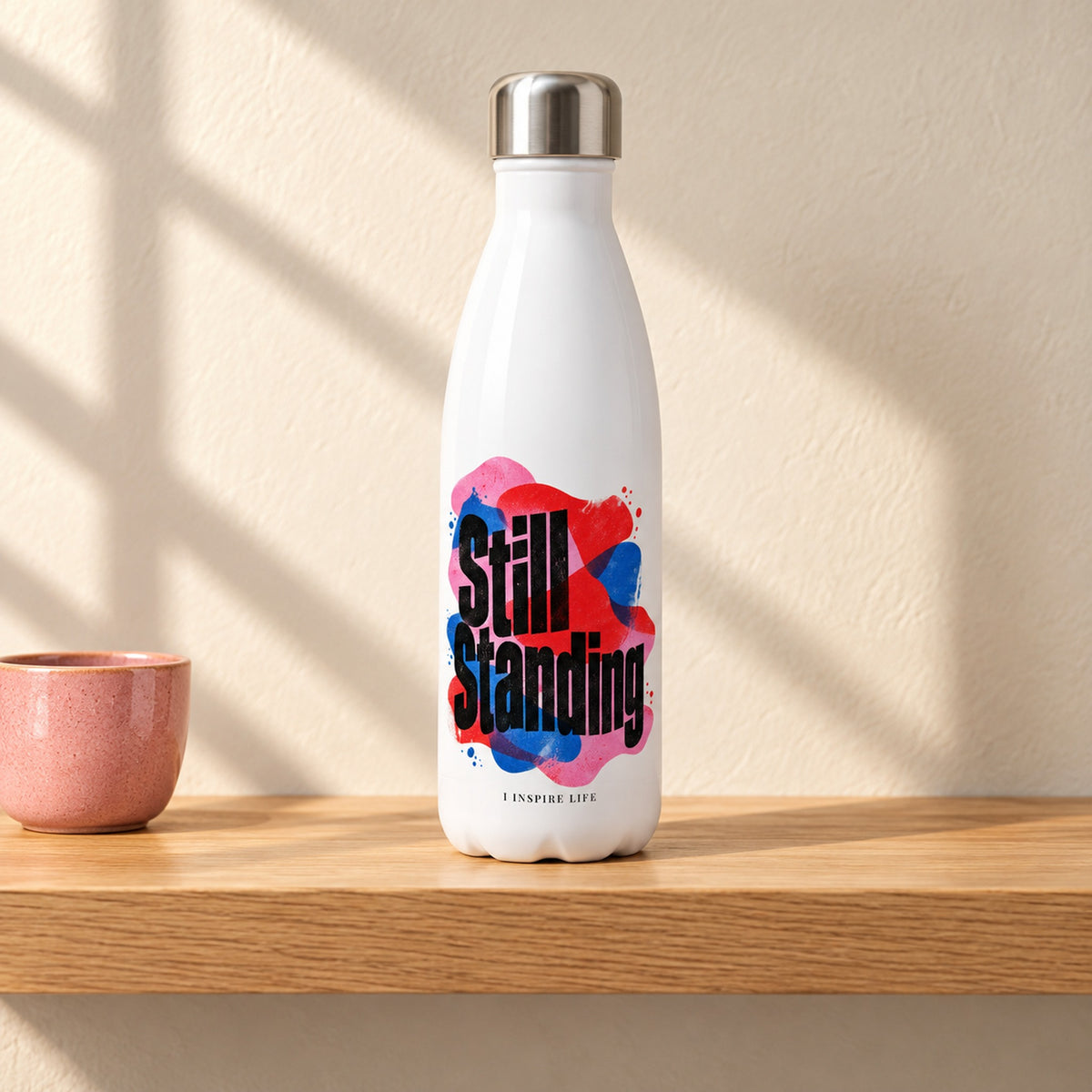 Still Standing 17 oz white stainless steel water bottle on a light oak shelf beside a small pink ceramic cup, design facing forward