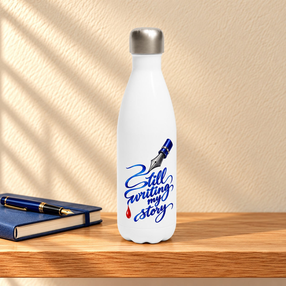 Still Writing My Story white 17 oz water bottle with a fountain pen nib and blue calligraphy, standing on a light oak shelf beside a notebook and pen