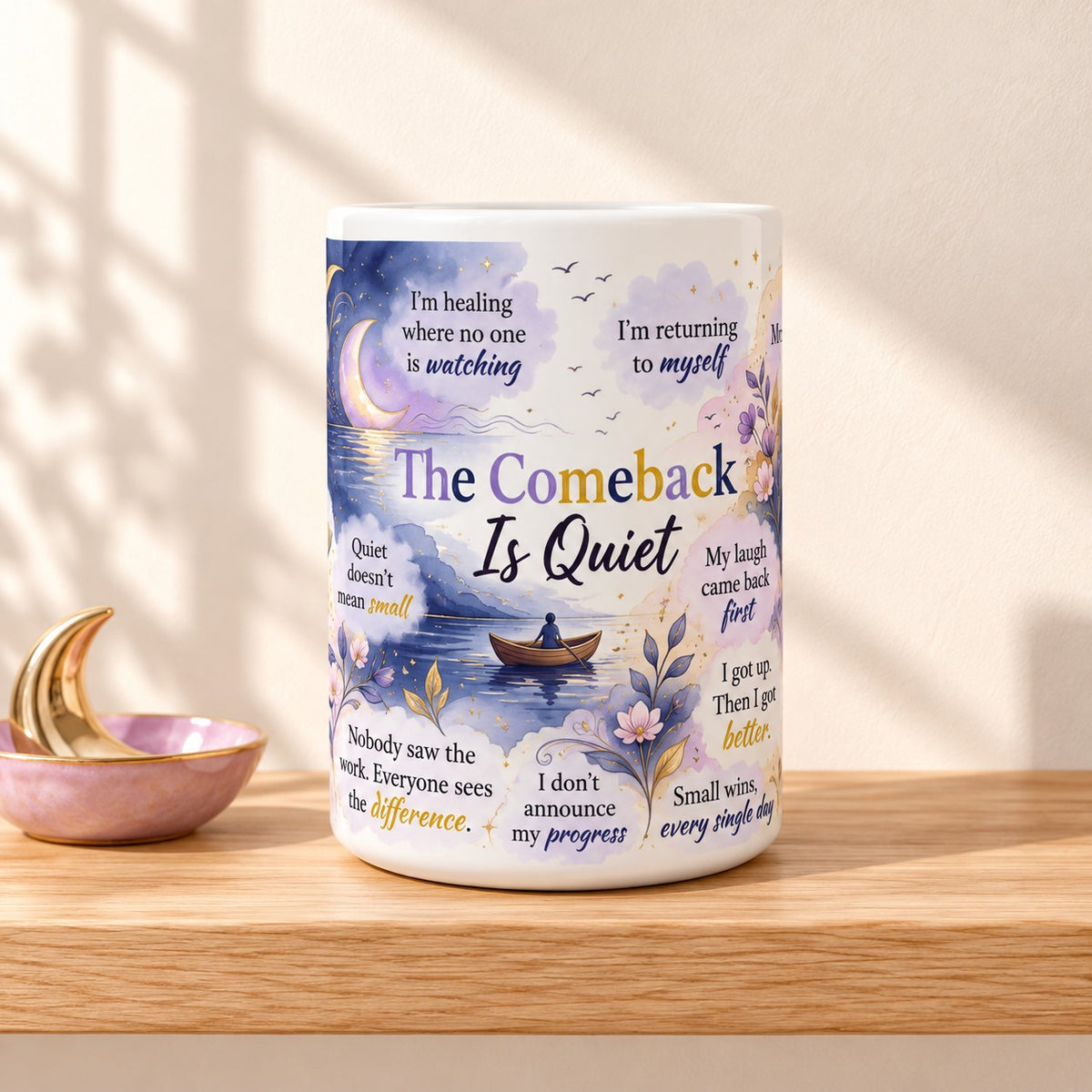 The Comeback Is Quiet 15 oz white mug with the title facing forward, resting on a light oak shelf beside a lavender dish with a gold moon