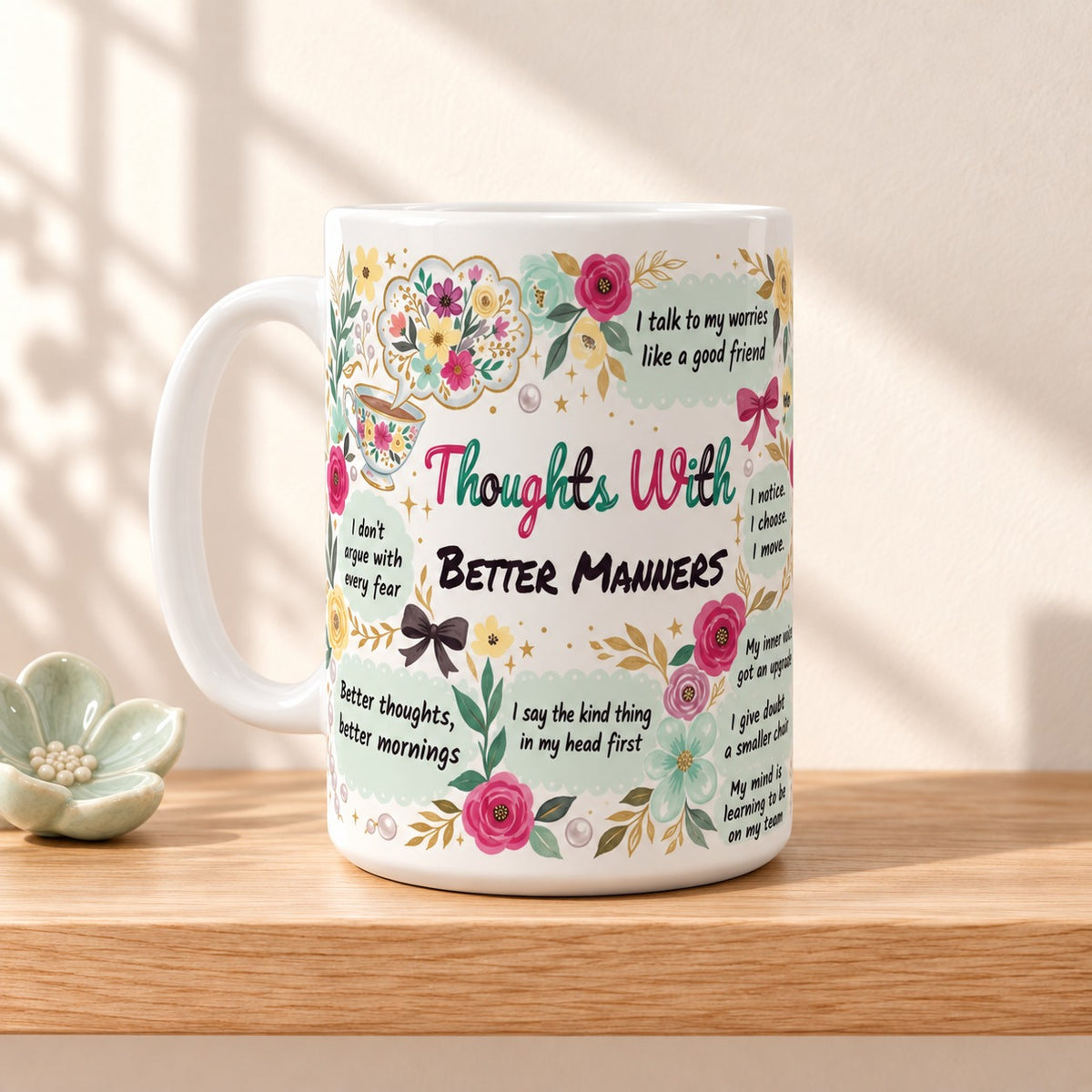 Thoughts With Better Manners 15 oz white mug with the title facing forward, resting on a light oak shelf beside a mint flower dish