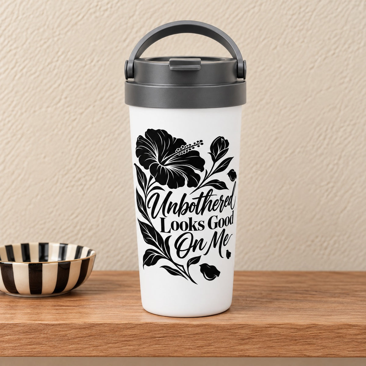 Unbothered Looks Good On Me 15 oz white travel mug with a bold black hibiscus and script lettering, standing on a light oak shelf beside a striped dish